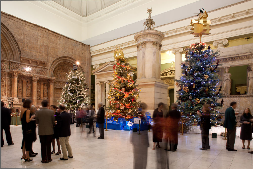 An image of Christmas trees in the Architecture Hall of Carnegie Museum of art. There are people mingling in the hall at a fundraiser. 