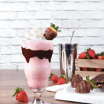 Chocolate-Dipped Strawberry Milkshake sits in a milkshake glass, pink in color. A strip of chocolate goes across the middle of the glass.