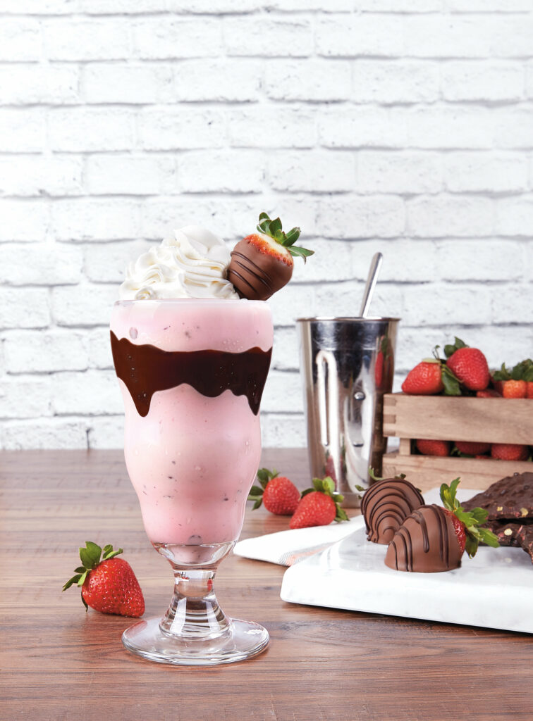Chocolate-Dipped Strawberry Milkshake sits in a milkshake glass, pink in color. A strip of chocolate goes across the middle of the glass.