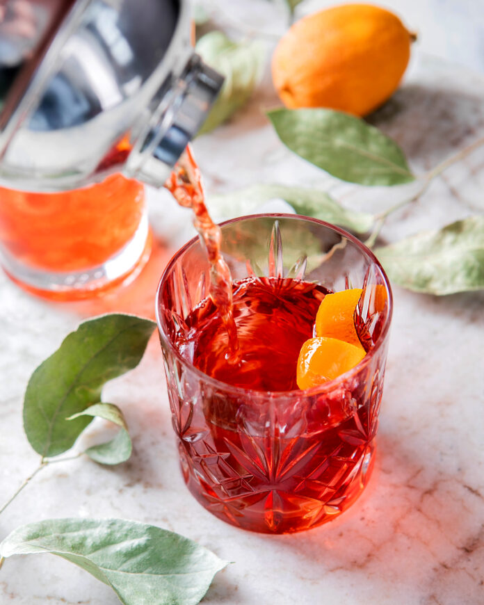 Red cocktail being poured into a clear glass from a silver cocktail shaker on a white table with leaves and citrus
