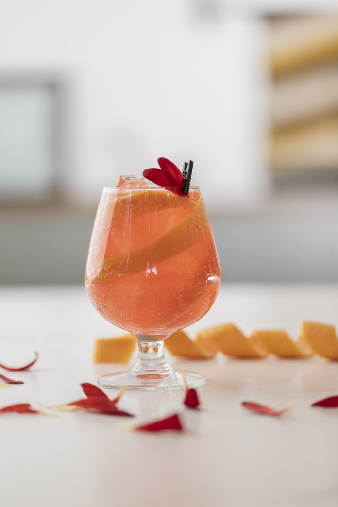 Orange cocktail sits in a glass surrounded by red flower petals and orange peel.