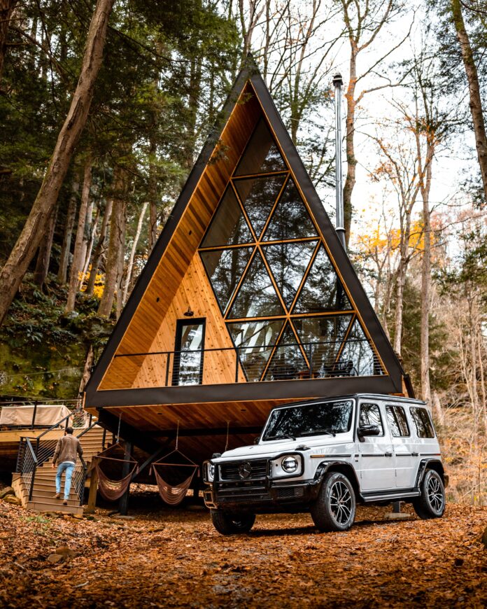 An A frame cabin with big glass windows in the woods. In front sits a grey/white Jeep to the right of the image. Long Weekend idea for Pittsburgh Travel
