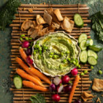 A flat-lay image of an Edamame and Kale hummus dish with carrots, cucumbers, and radishes surrounding the plate.