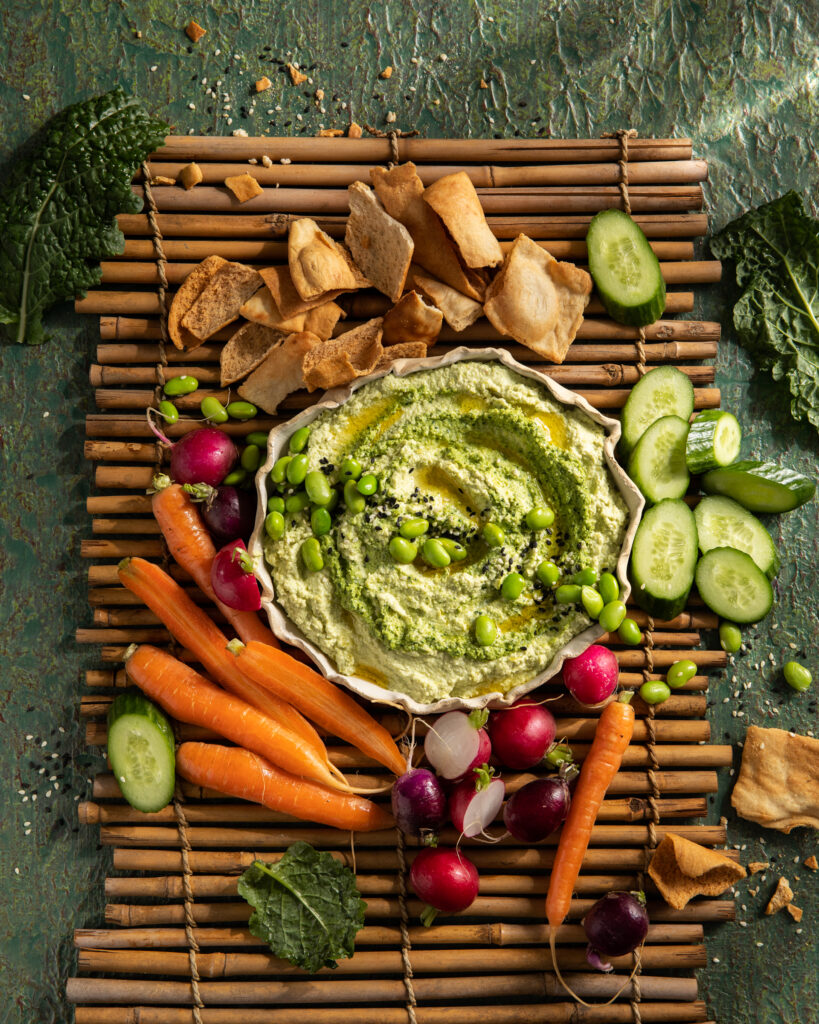 A flat-lay image of an Edamame and Kale hummus dish with carrots, cucumbers, and radishes surrounding the plate.