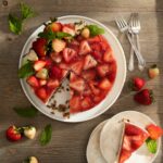 A plate of Strawberry Gelatin Pretzel Cake on a wooden surface with three forks and fresh strawberries lying on the side. A piece of cake on a smaller plate that's stacked over two empty plates.