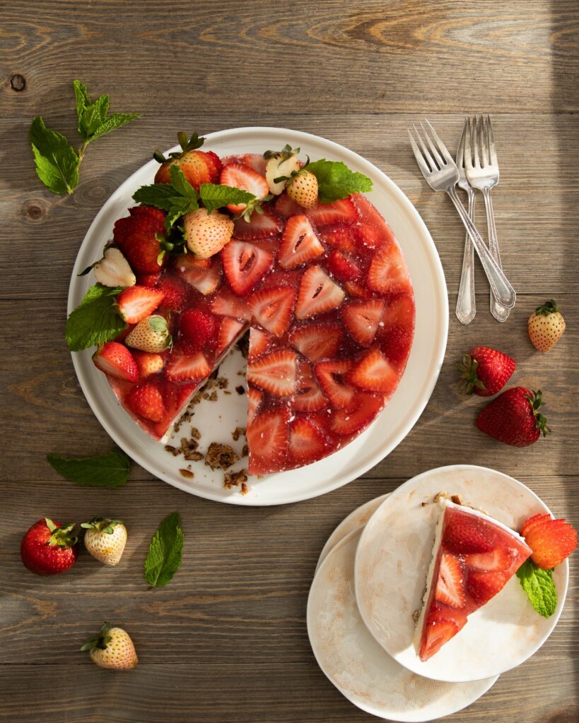 A plate of Strawberry Gelatin Pretzel Cake on a wooden surface with three forks and fresh strawberries lying on the side. A piece of cake on a smaller plate that's stacked over two empty plates.