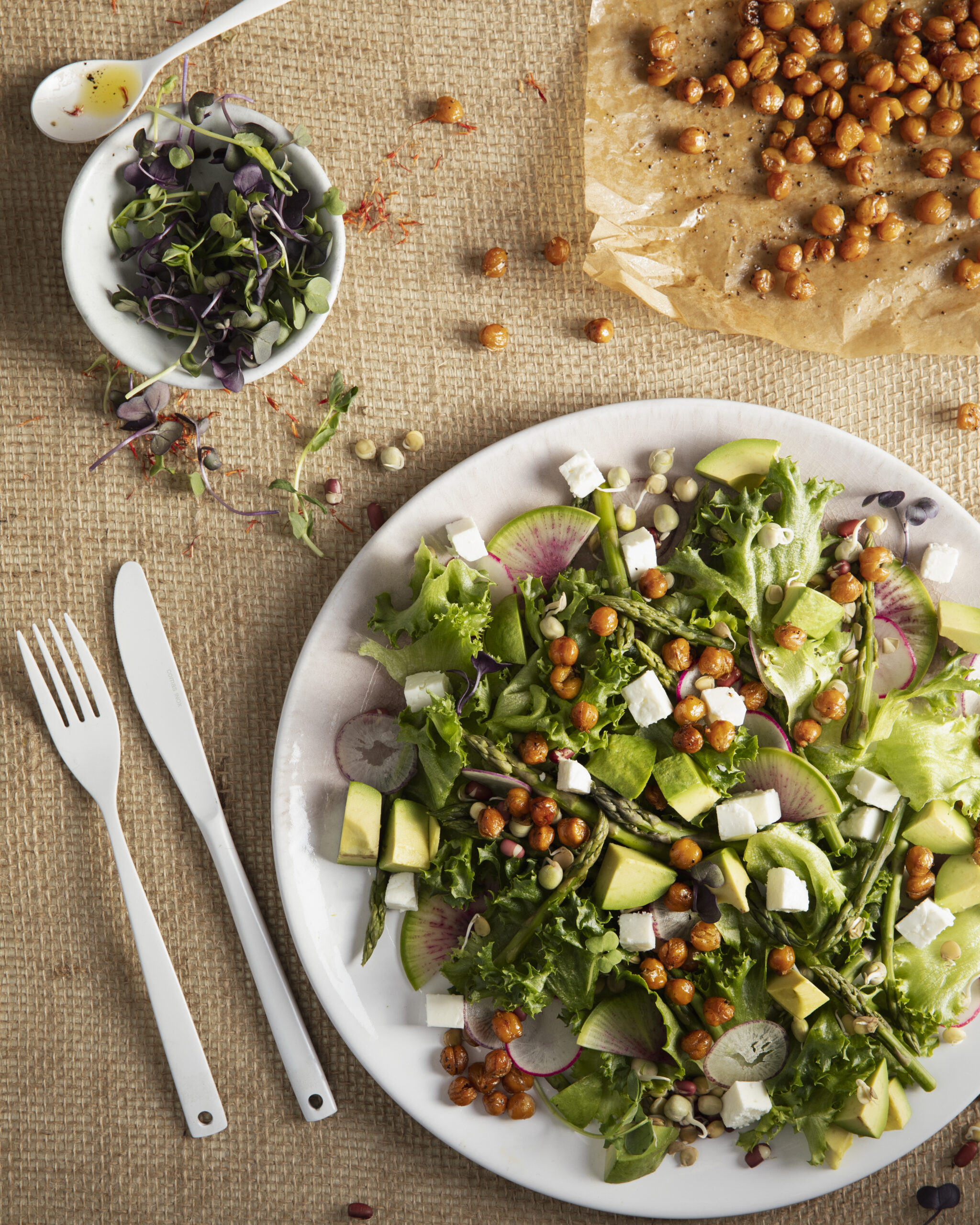 An aerial view of a Spring Time Salad with Saffron Vinaigrette, which sits on a white plate. A white fork and knife are positioned to the left of the salad.