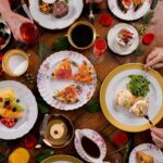 An overhead view of a wooden table set for brunch with plates of eggs benedict, steak, salmon flatbread, and fresh fruit salad.