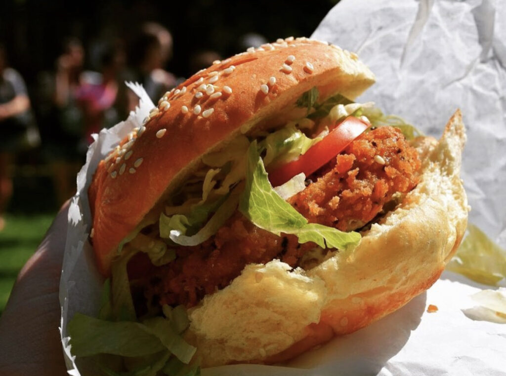 a vegan sandwich that looks like fried chicken with lettuce and tomatoes between 2 burger buns, meatless veggie dish