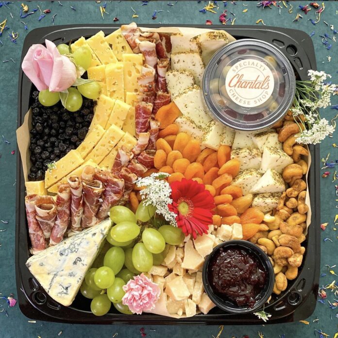 a variety of meats cheeses nuts fruit and spreads on a square disposable board decorated with flowers on a teal background