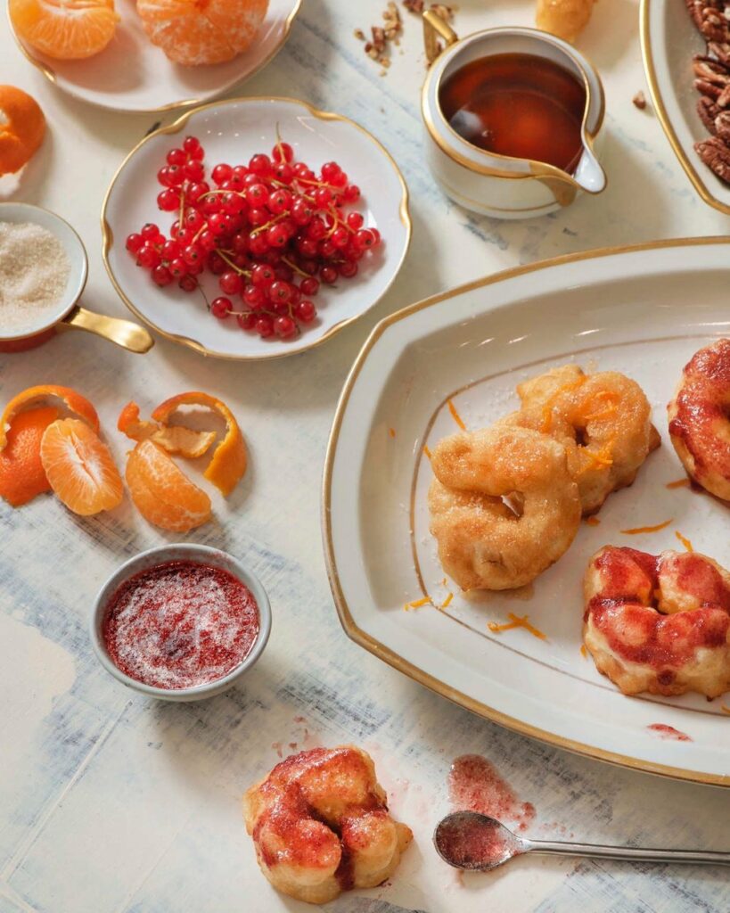 Isfeng Andalusian Donuts sit on a plate to the right side of the frame. Oranges and cherries sit to the left.