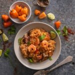 Jambalaya in a bowl placed on a ceramic surface along with a spoon, spices, and lemon