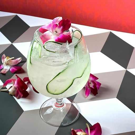 cocktail in a clear glass with a cucumber ribbon and an orchid on a black and white 3d cube print table with more orchids around the glass