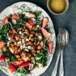 A Strawberry Kale Salad sits on a white plate featuring small green patterns. A silver fork and spoon sit to the right of the plate.