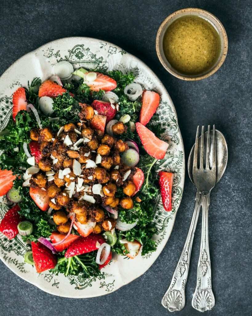 A Strawberry Kale Salad sits on a white plate featuring small green patterns. A silver fork and spoon sit to the right of the plate.