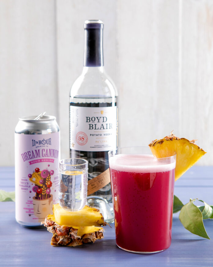 a colorful can of a sour beer to the left of a bottle of vodka both sitting behind a shot glass and a sour brew garnished with pineapple