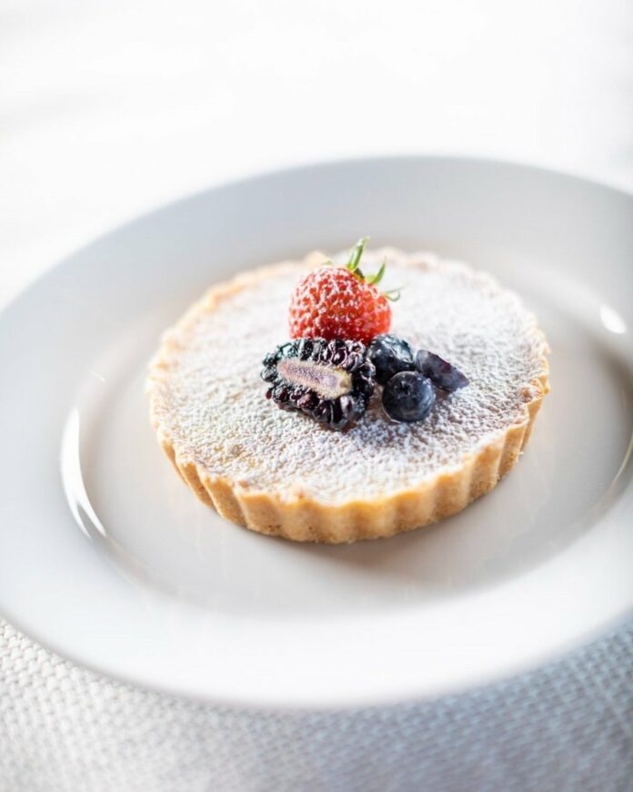 A lemon tart topped with berries and powdered sugar, placed on top of a white plate.