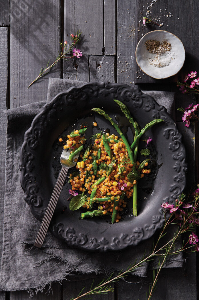 Lentil-Asparagus-Corn Salad Orange lentil Asparagus Corn Salad sits on a black plate on top of a black wooden table.