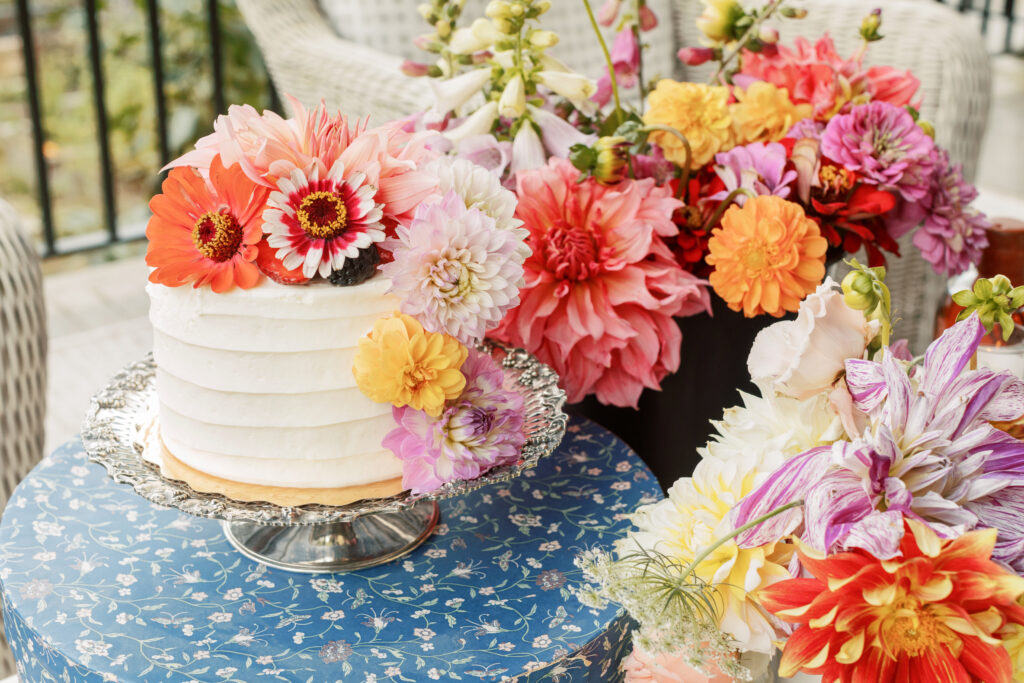 round cake on a glass cake plate with a vast variety of pastel colored Dalias and zinnias on a round table with a blue tablecloth 