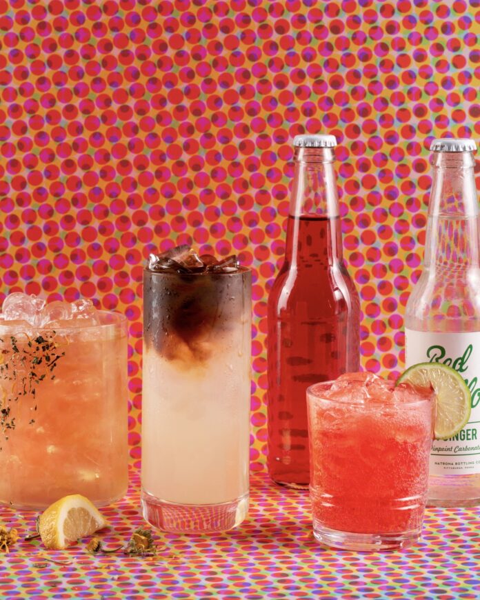 3 iced beverages and 2 glass bottled sodas in front of a brightly colored polka dot background