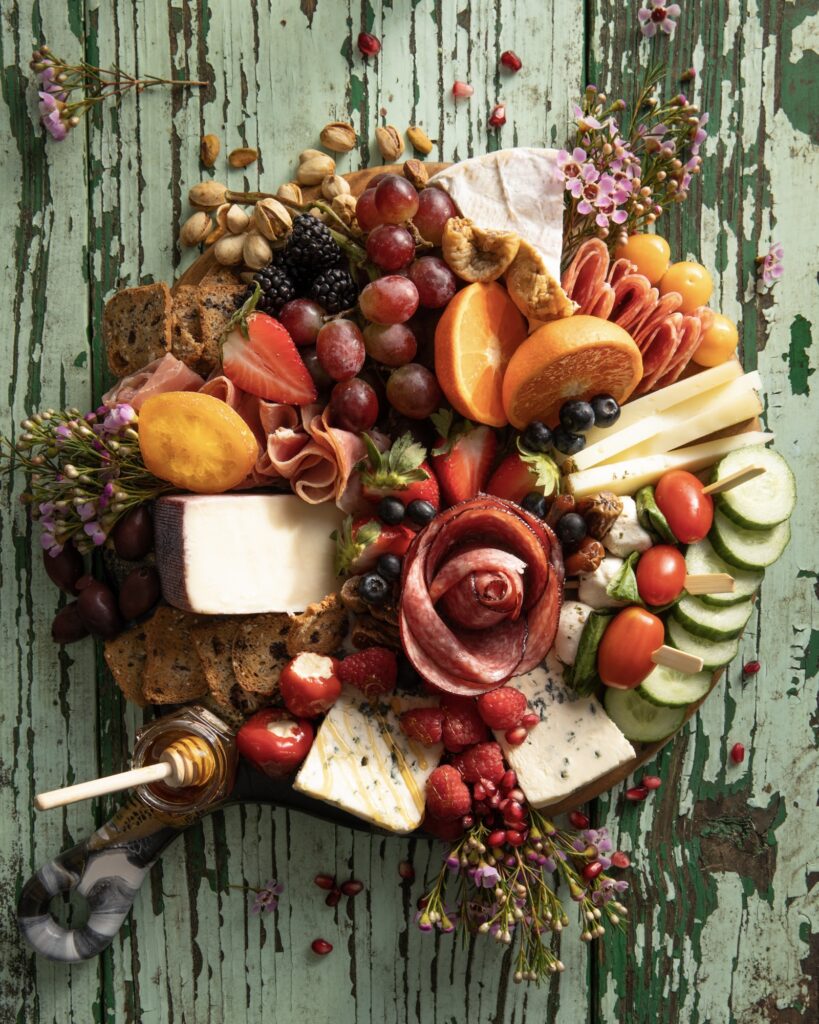 a round board with meats cheeses fruits vegetables and crackers on a rustic green painted board