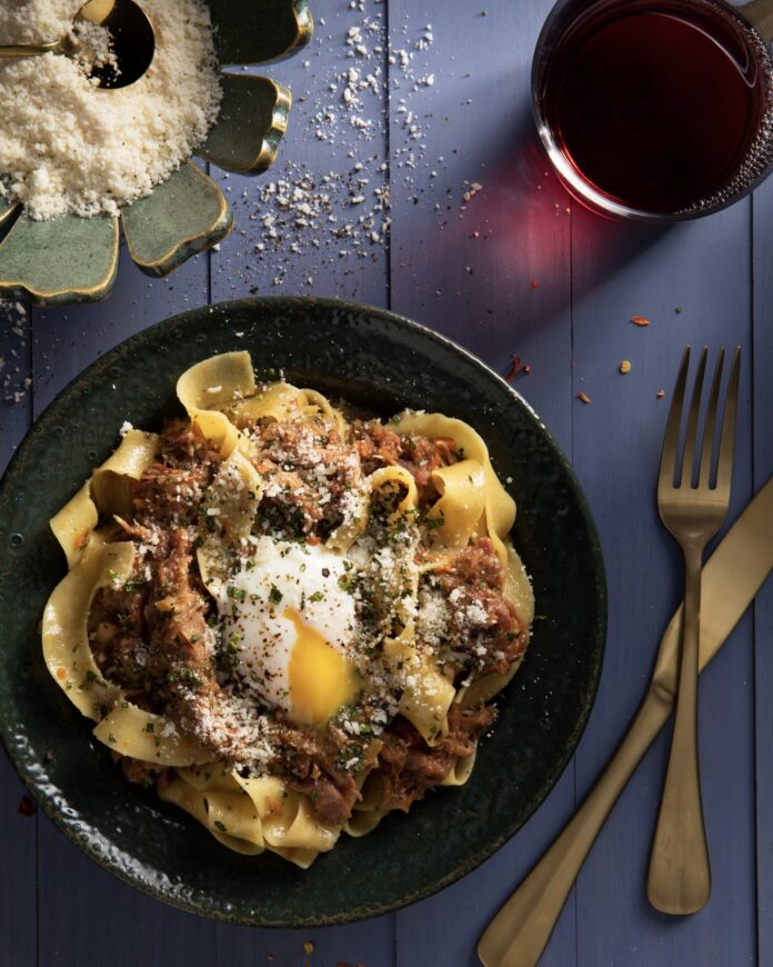 a wide black bowl filled with tagliatelle pasta, a meaty sauce and a broken open soft cooked egg with a loose yolk