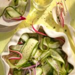 a bowl filled with Nana’s Cucumber Salad next to a smaller bowl and a fork and a knife