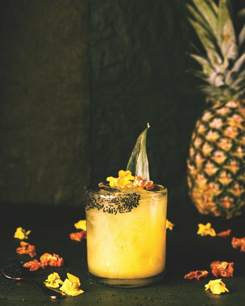 a yellow glass of Pineapple Jalapeño Margarita topped with a pineapple leaf, flowers, and black salt on the rim.