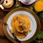 Pumpkin Pancakes topped with Apple Compote served on a plate with a fork with butter, syrup, and a cup of juice on the sides