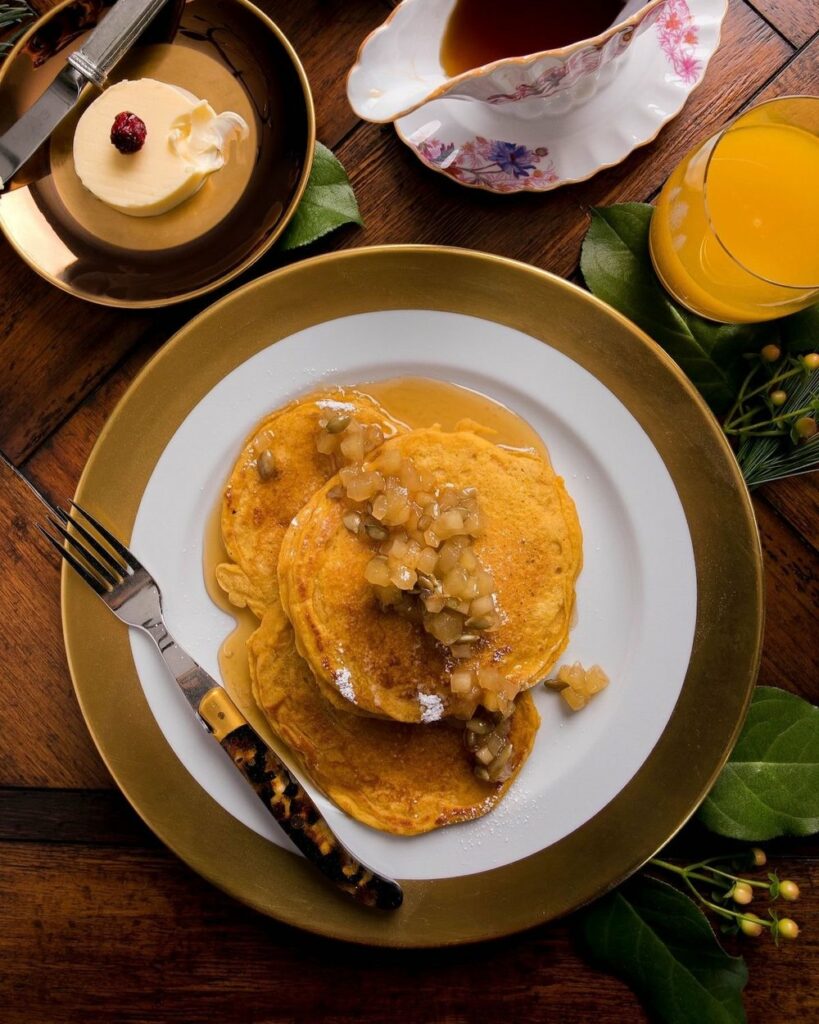 Pumpkin Pancakes topped with Apple Compote served on a plate with a fork with butter, syrup, and a cup of juice on the sides