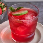 Red Summer Punch sits in a small drinking glass on a small white plate, garnished with lime.