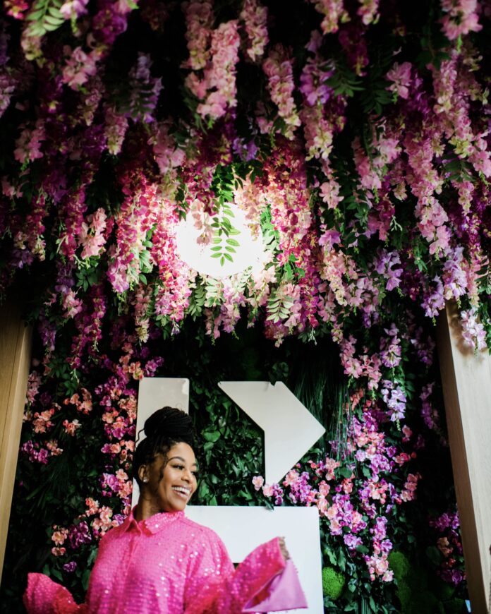 a woman of color in a bright pink dress posing under a ceiling of hanging pink and purple wisteria. she's at a pittsburgh restaurant with ambience.