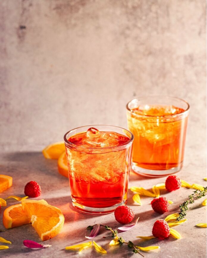 Two Sandia Sunset Campari cocktails sit in rocks glasses, one slightly in front of the other. Orange and red in color, raspberries and oranges sit on the table around the cocktails.