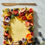 A rectangular seasonal fruit tart with creamy pastry cream filling, elaborately garnished with fresh berries, sliced peaches, figs, and edible flowers on a white marble surface.