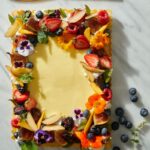 A rectangular seasonal fruit tart with creamy pastry cream filling, elaborately garnished with fresh berries, sliced peaches, figs, and edible flowers on a white marble surface.