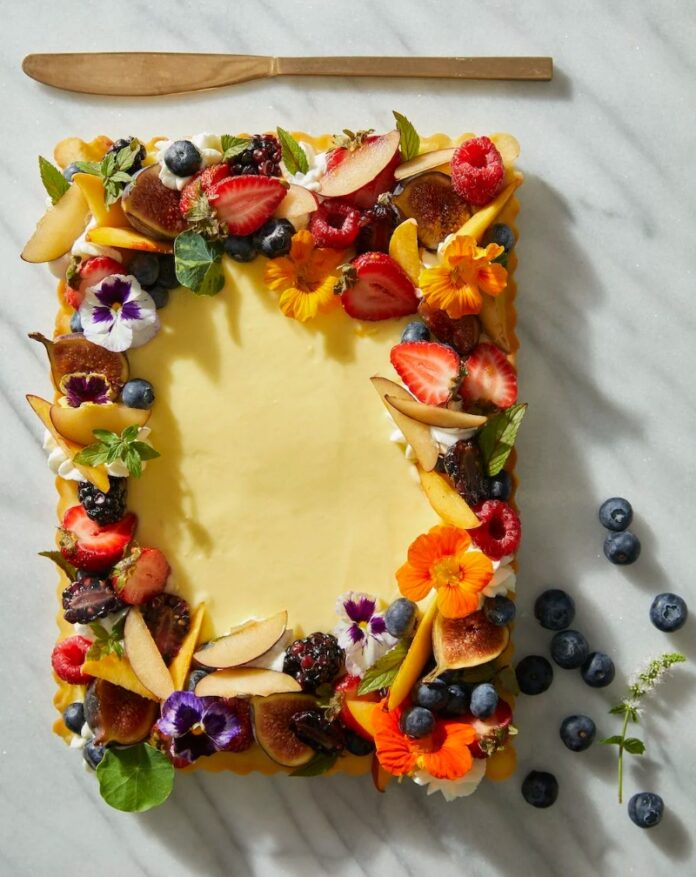 A rectangular seasonal fruit tart with creamy pastry cream filling, elaborately garnished with fresh berries, sliced peaches, figs, and edible flowers on a white marble surface.