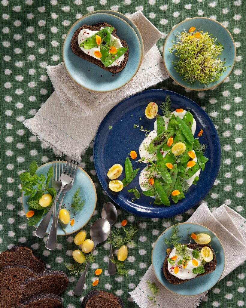 Dew peas in a dark blue plate with four light blue plates surrounding it. Other items are napkins, pieces of bread, forks, and spoons.