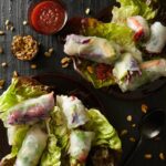 A flat lay of fresh spring rolls with a sweet potato noodle filling, arranged on lettuce leaves with a side of dipping sauce and roasted peanuts.