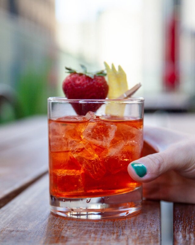 A Strawberry Negroni Bianco sits in a rocks glass, garnished with a strawberry and twist of a peel of lemon. A hand holds the glass on the right.