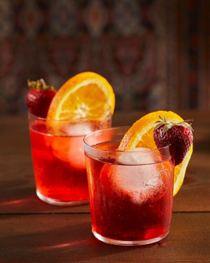 Two Strawberry Rhubarb Negronis sit in short glasses, one slightly in front of the other. Deep red in color, they are garnished with a strawberry and orange slice.