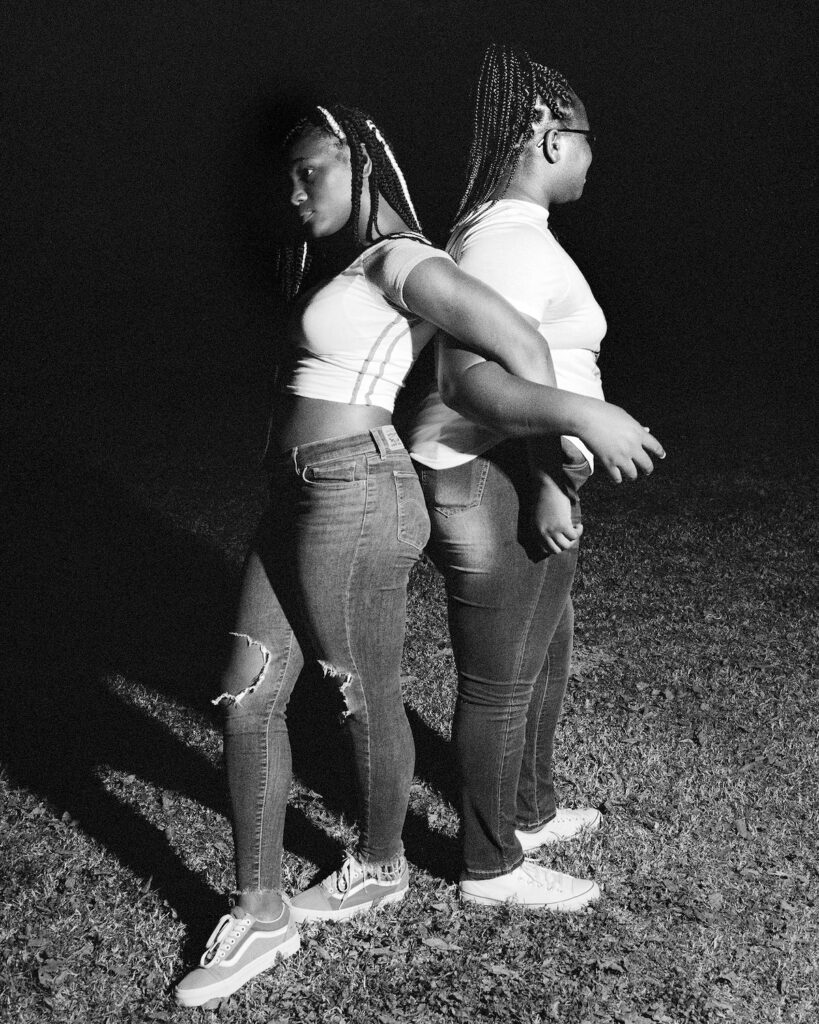 Two teenage Black girls stand back to back with their arms interlinked. The image is in black and white.