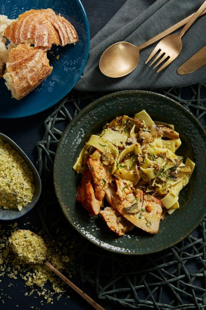 A dark green bowl of tuna confit casserole with pappardelle pasta and mushrooms, served with crusty bread and a side of herb breadcrumbs, and brass cutlery.