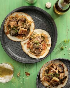 Three pork belly soft tortilla tacos, two on a tray and one on a tray a bottle of beer and a glass of beer on a green table