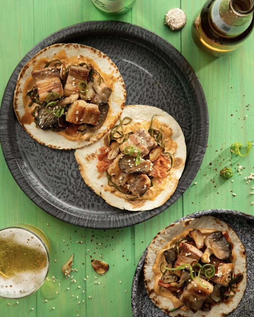 Three pork belly soft tortilla tacos, two on a tray and one on a tray a bottle of beer and a glass of beer on a green table