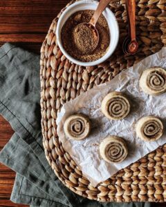 An aerial view of five cinnamon rolls on a piece of parchment paper next to a bowl of cinnamon with a spoon in it.