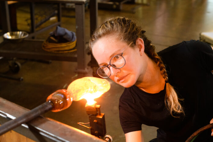 A white woman with french braids and glasses concentrates on blowing glass.