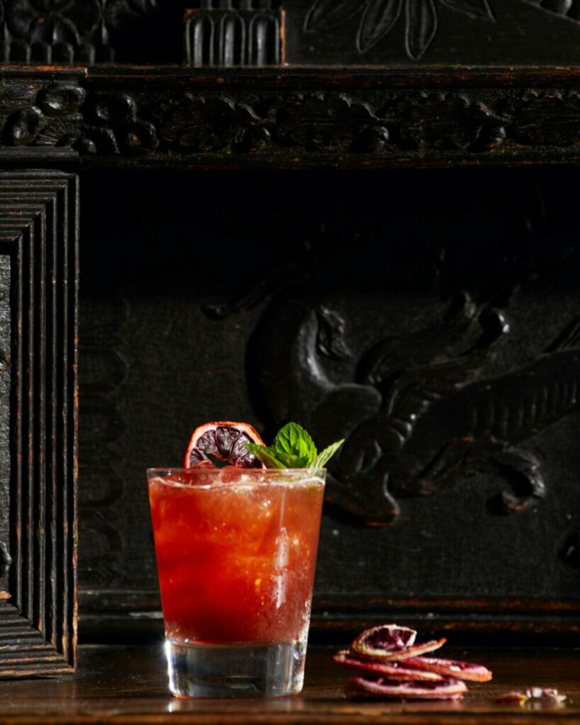 A strawberry fields cocktails sits in a rocks glass garnished with dehydrated blood orange slice and mint.