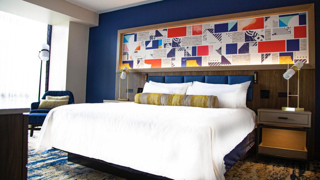 a hotel room with a large window and bed with white bedding and a colorful large framed geometric piece above the bed