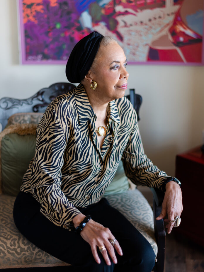 Jo-Ann Bates ©JOEYKENNEDY AWC HR D-0002 Jo-Ann Bates, a Black female artist, sits on the side of a chair and looks to the right. She wears a small black hat on her head and a zebra print button up.