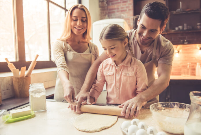 Cute little girl and her beautiful parents in aprons are flattening dough and smiling while baking in kitchen at home and nurturing for the future. Recipes for Financial Success Summer 2023